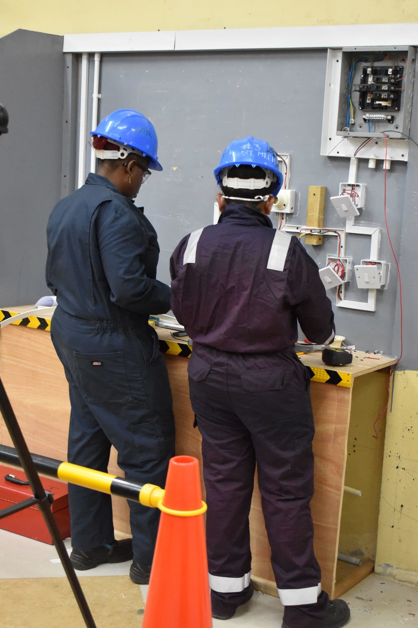 Two people in coveralls and safety helmets at an electrical training panel