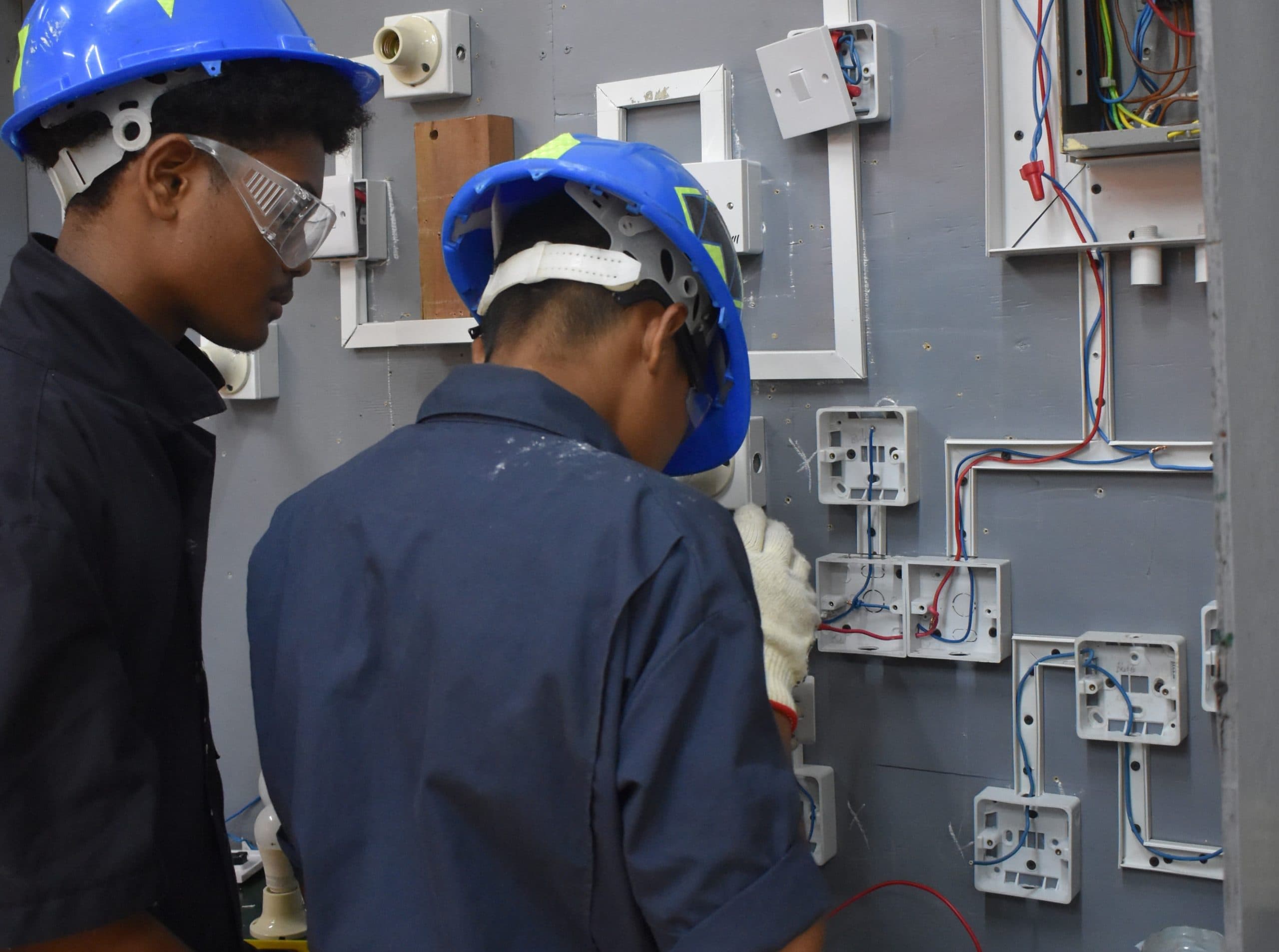 Two learners in safety helmets working at an electrical training panel